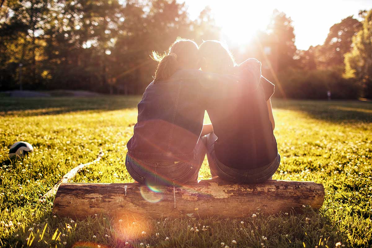 Två unga tjejer sitter på en stock i solnedgångnen och kramar om varandra
